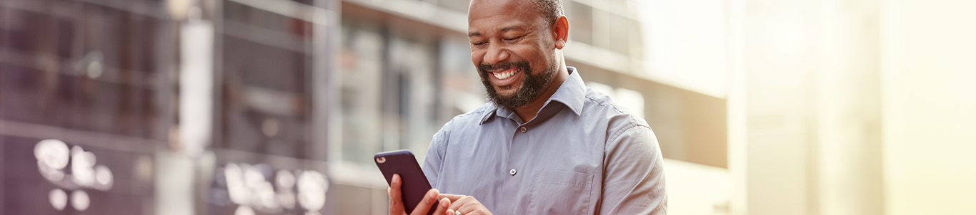 A man smiling while reading something on his phone, standing outside an office building